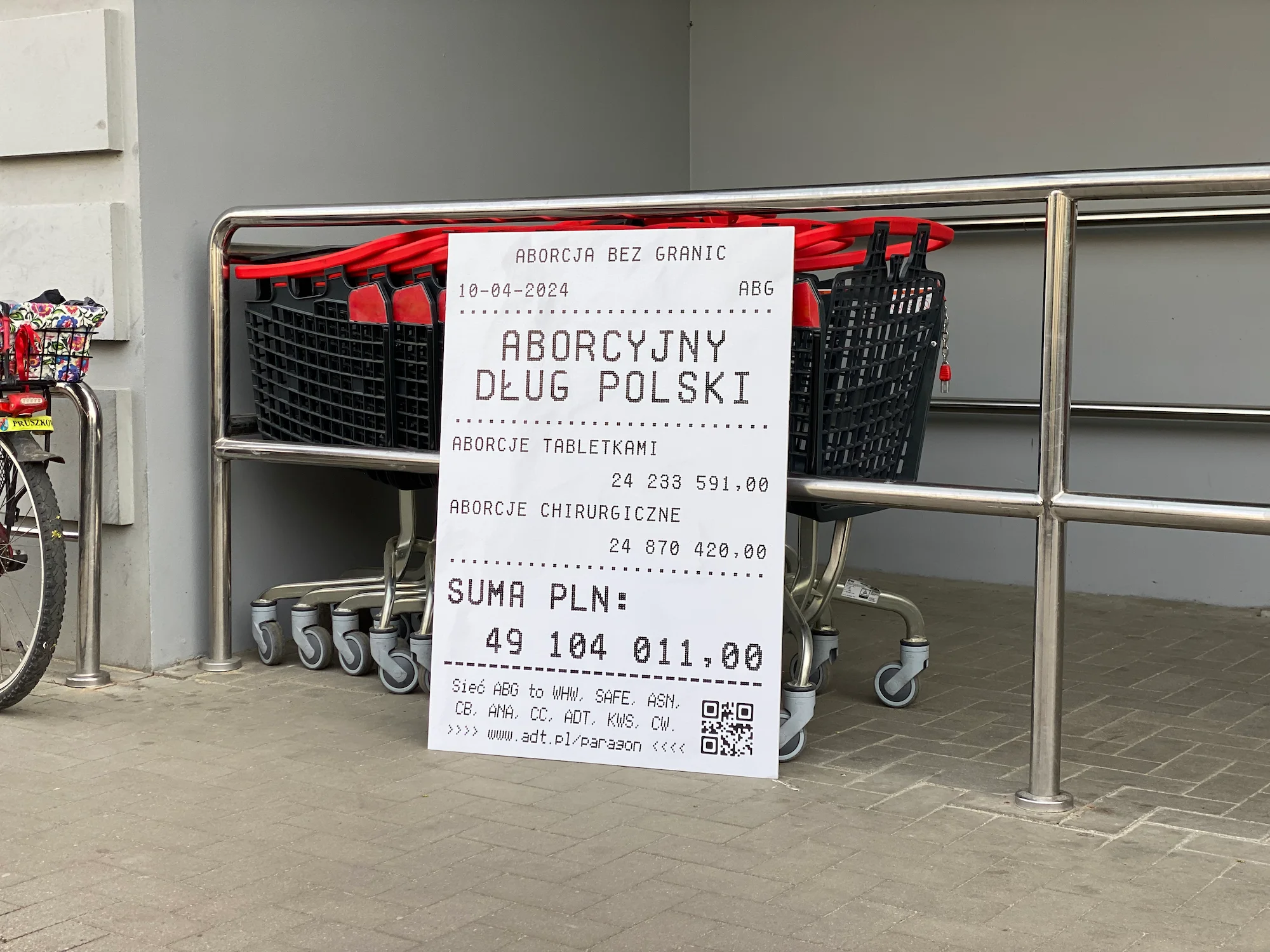 Paragon z Polskim Długiem Aborcyjnym oparty o barierki przy wózkach zakupowych przed sklepem.